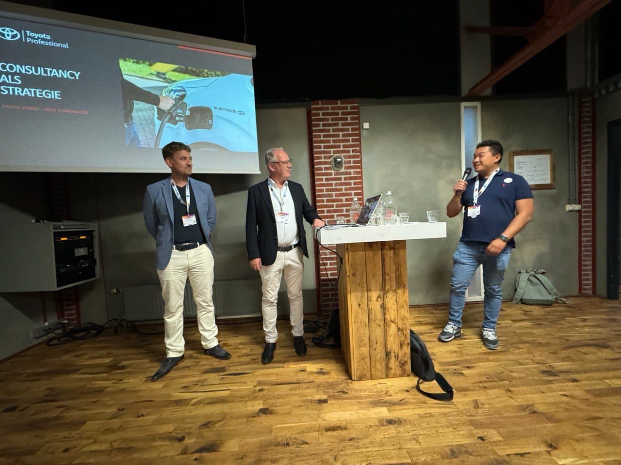 Wing Cheung spreekt met een microfoon op een podium, naast twee andere sprekers